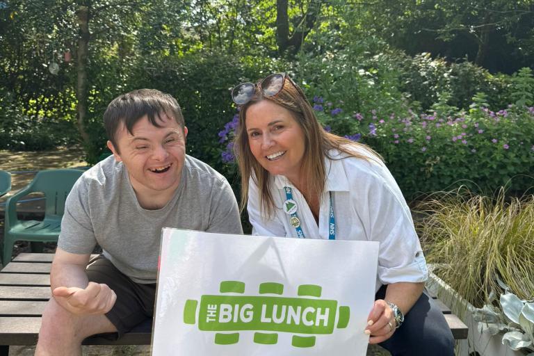 Two people sat on a bench, smiling and holding up a sign saying "the big lunch"